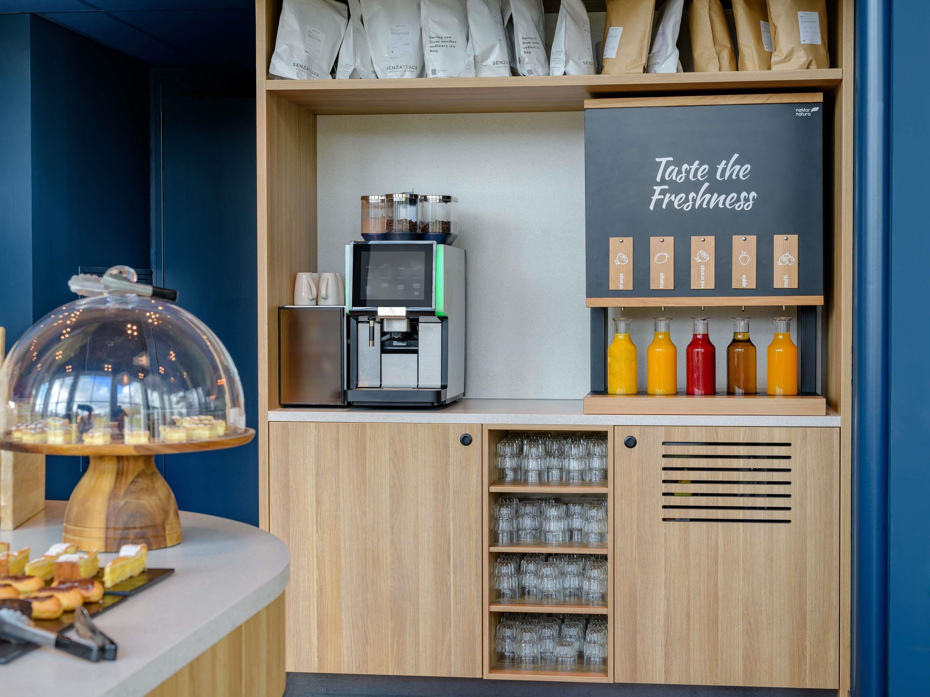 A coffee station with a coffee machine, a glass display case with pastries, and a sign that says 'Taste the Freshness.'