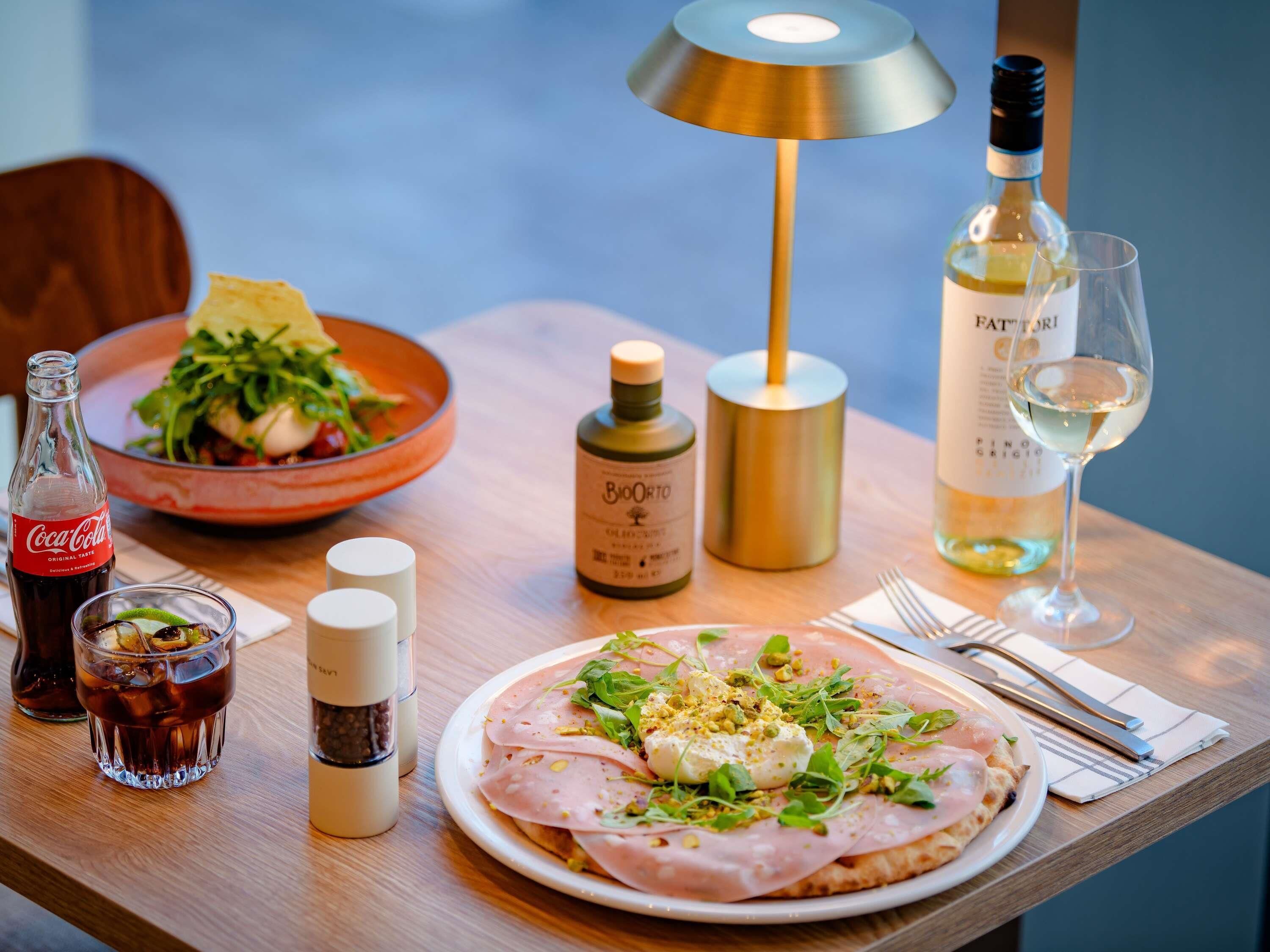 Una mesa con un plato de comida, una botella de vino, una botella de aceite de oliva, una botella de Coca-Cola, un vaso de Coca-Cola, un vaso de vino y un salero y pimentero.