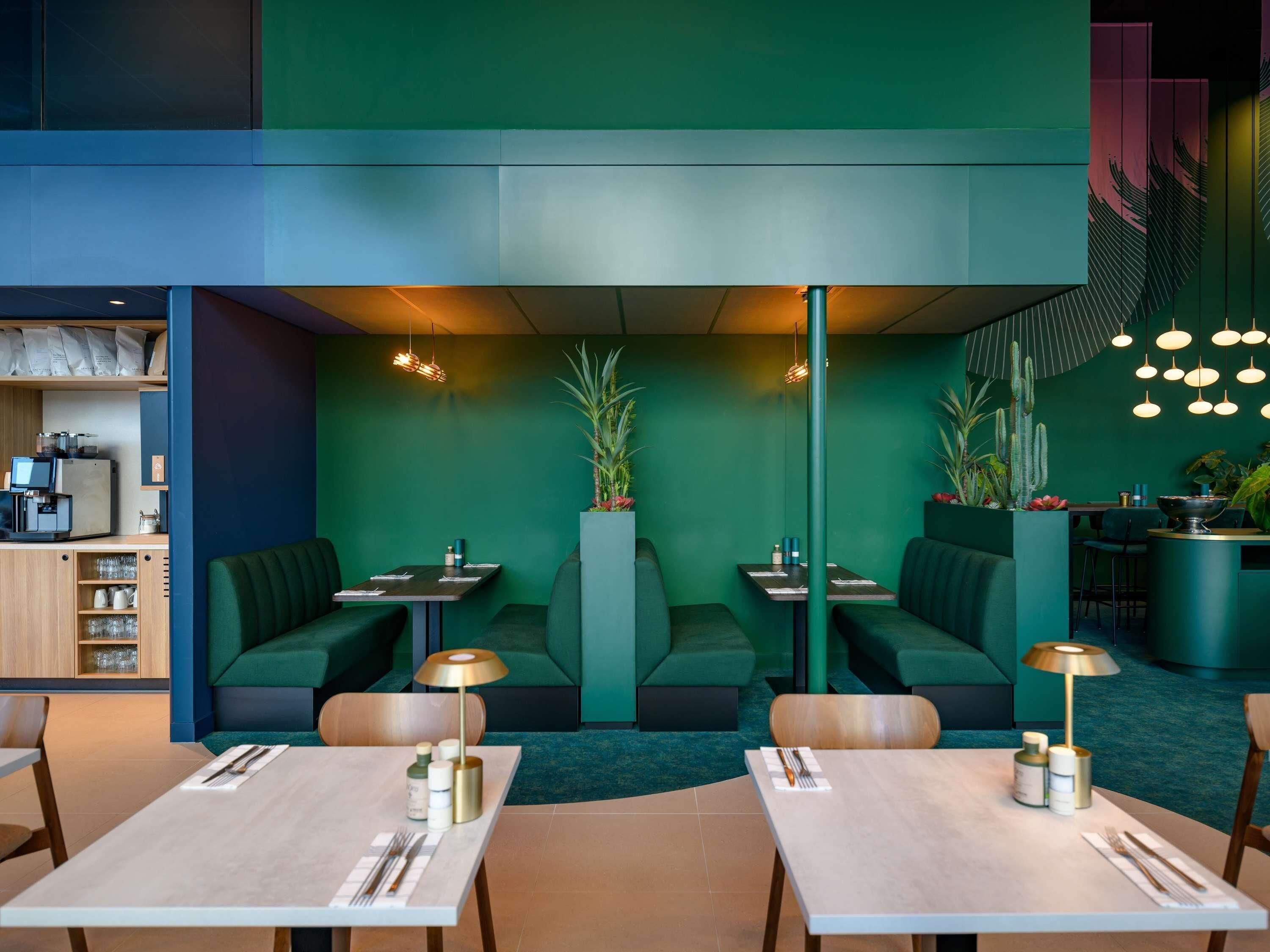 A restaurant with green walls, multiple seating areas including booths and tables, and a bar area with plants.