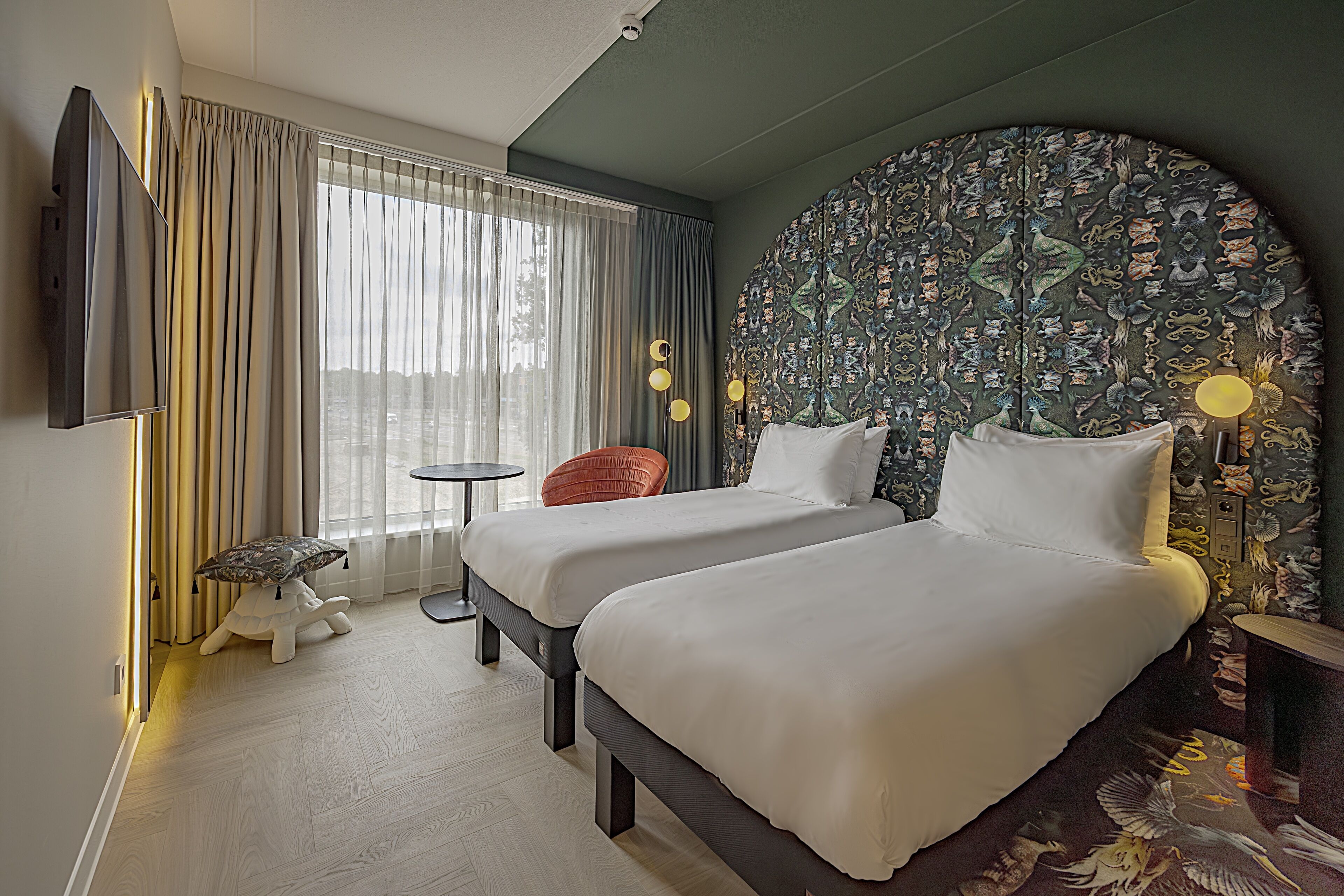 A hotel room with two beds, a TV, a small table, a chair, and a large decorative wall.