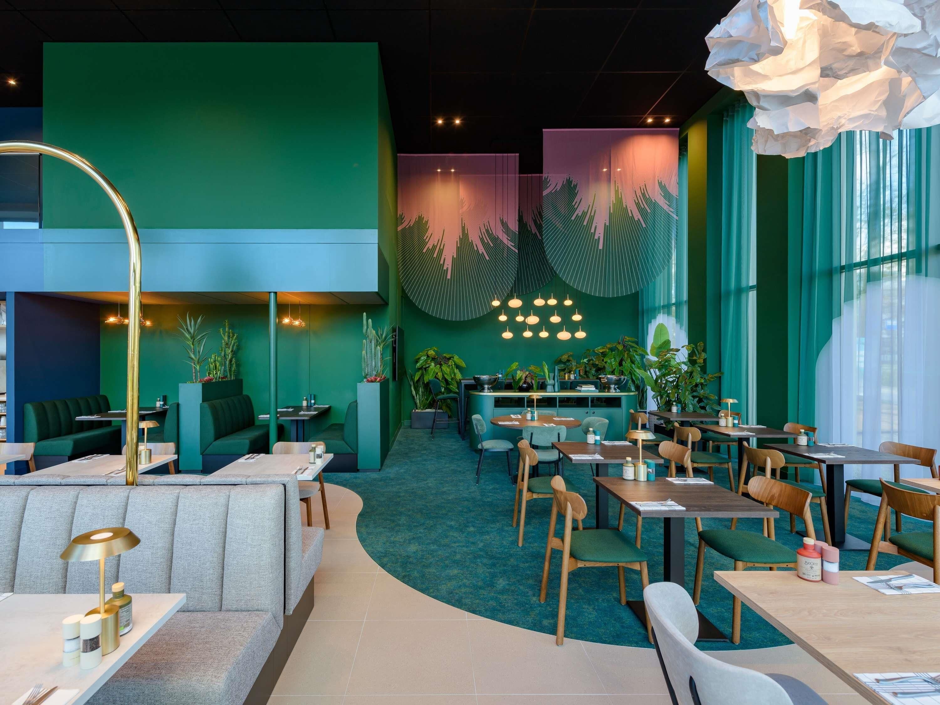 Een modern restaurantinterieur met groene wanden, meerdere zithoeken en decoratieve plafondlampen.