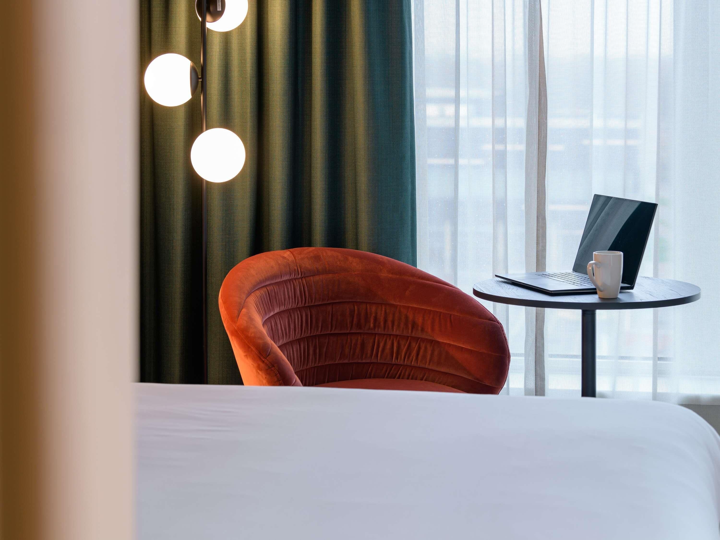 Een hotelkamer met een bed, een rode stoel, een klein rond tafeltje met een laptop en een beker, en een raam met gordijnen.