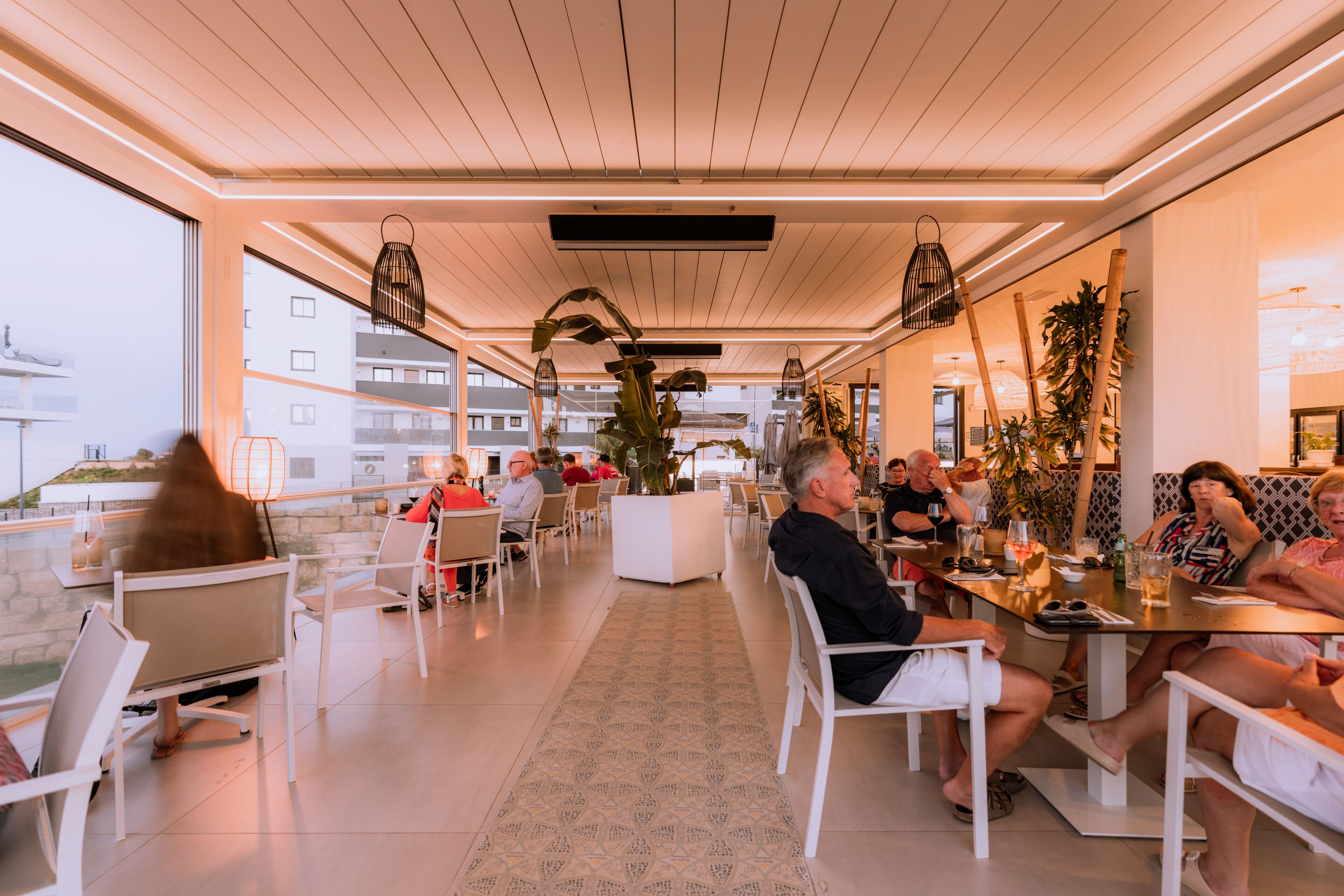 A modern indoor dining area with white furniture, large windows, and indoor plants.