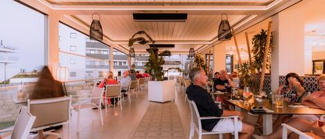 A modern indoor dining area with white furniture, large windows, and indoor plants.