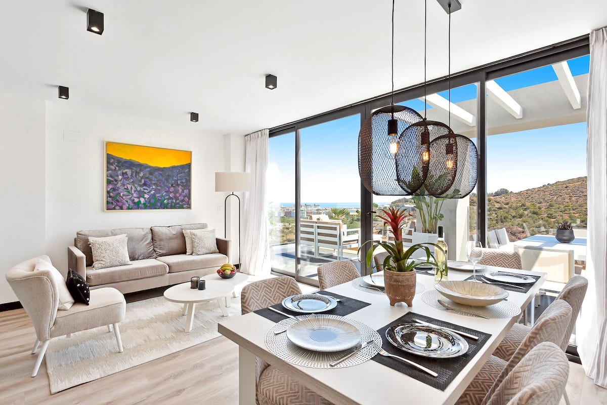 A modern living room with a sofa, coffee table, and dining area with a table set for two. Large windows offer a view of the city and hills.