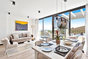 Un salon moderne avec un canapé, une table basse et un coin repas avec une table dressée pour deux personnes. De grandes fenêtres offrent une vue sur la ville et les collines.