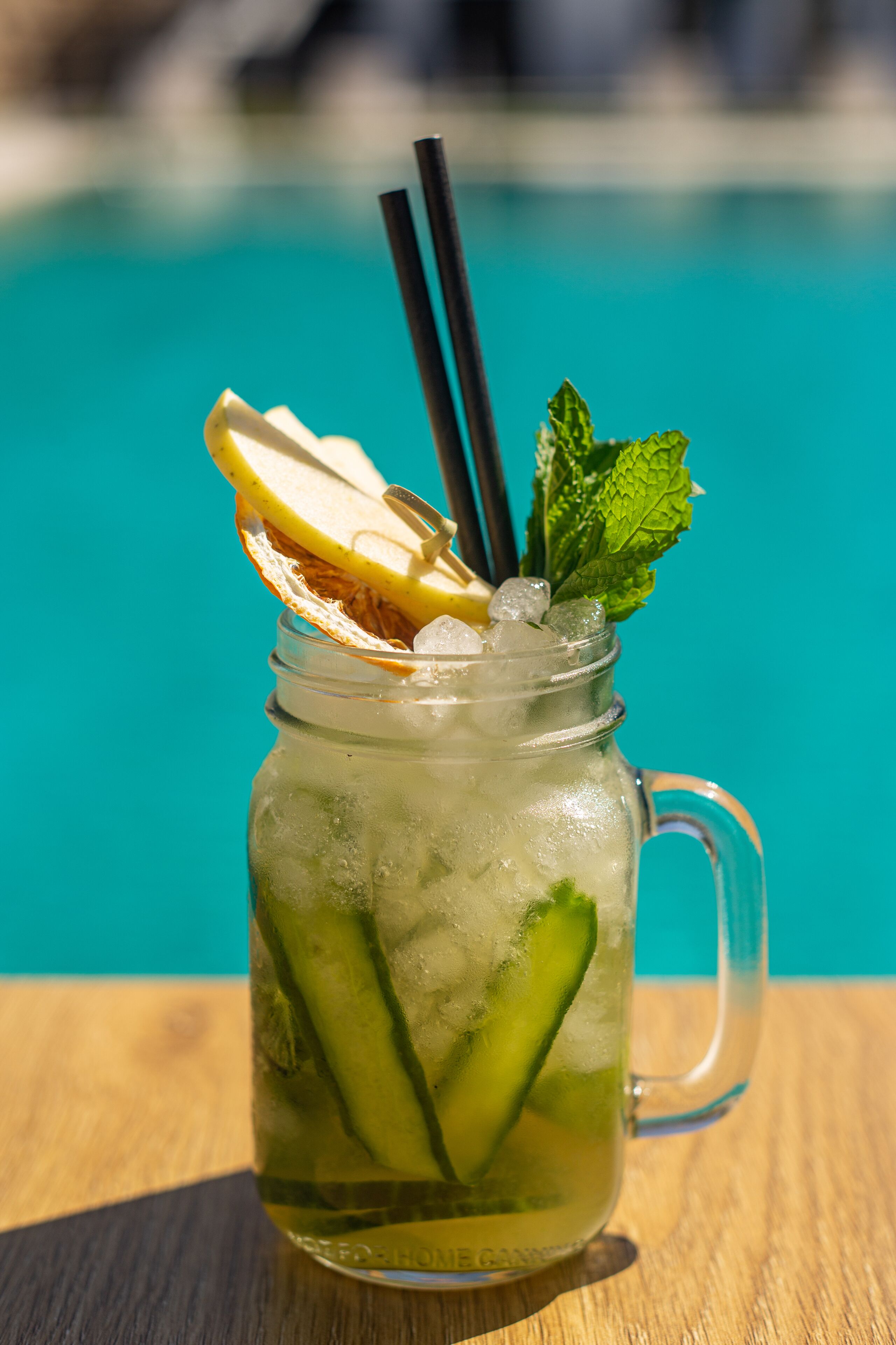 Una taza de vidrio transparente con asa, que contiene una bebida con cubitos de hielo, rodajas de limón y hojas de menta, sobre una superficie de madera con una piscina al fondo.