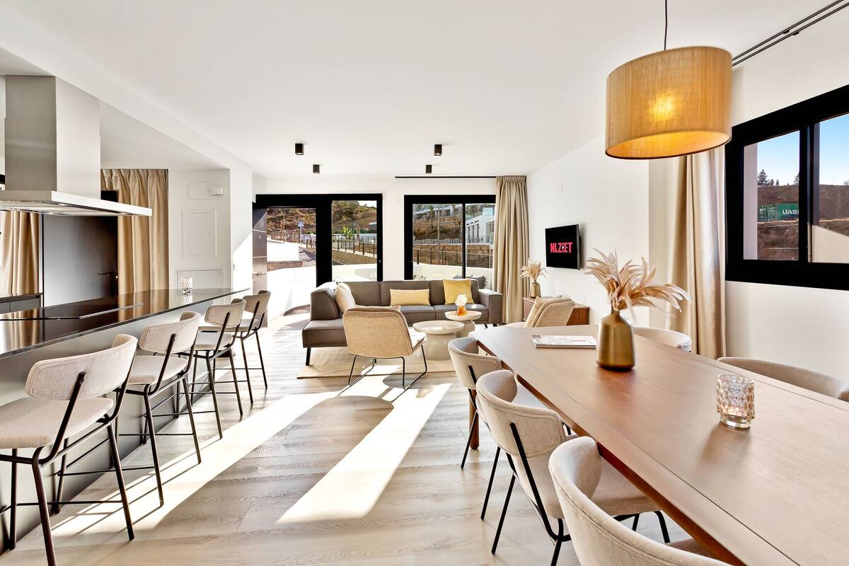 A modern living room with a sofa, coffee table, and dining area. There is a TV mounted on the wall and a window with a view of the outdoors.