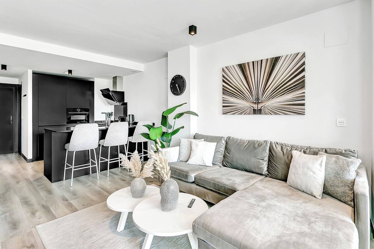 A modern living room with a grey sectional sofa, two white round tables, and a dining area with a kitchen in the background.