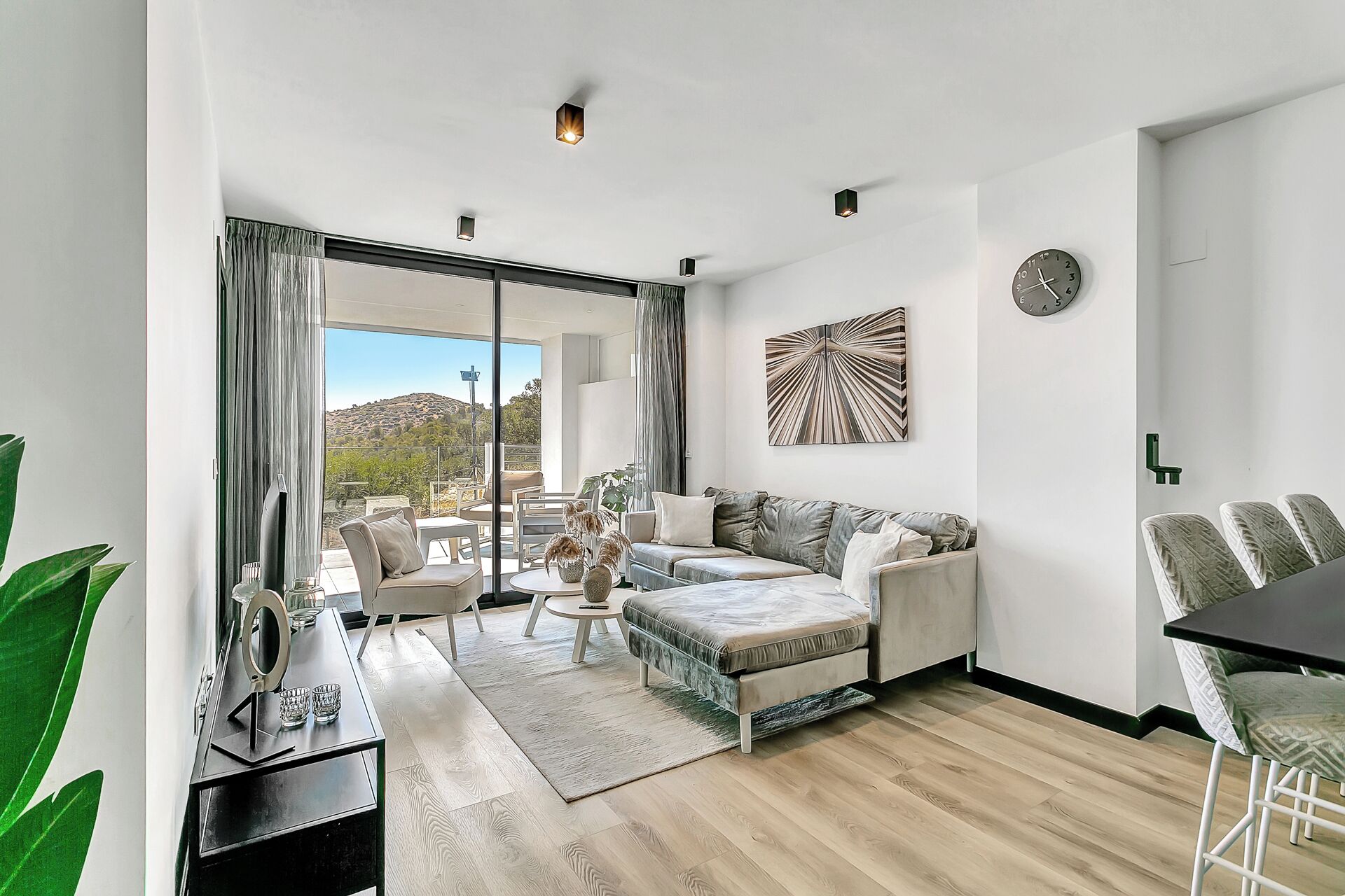 A modern living room with a sofa, coffee table, and chairs. There is a large window with a view of a hill.
