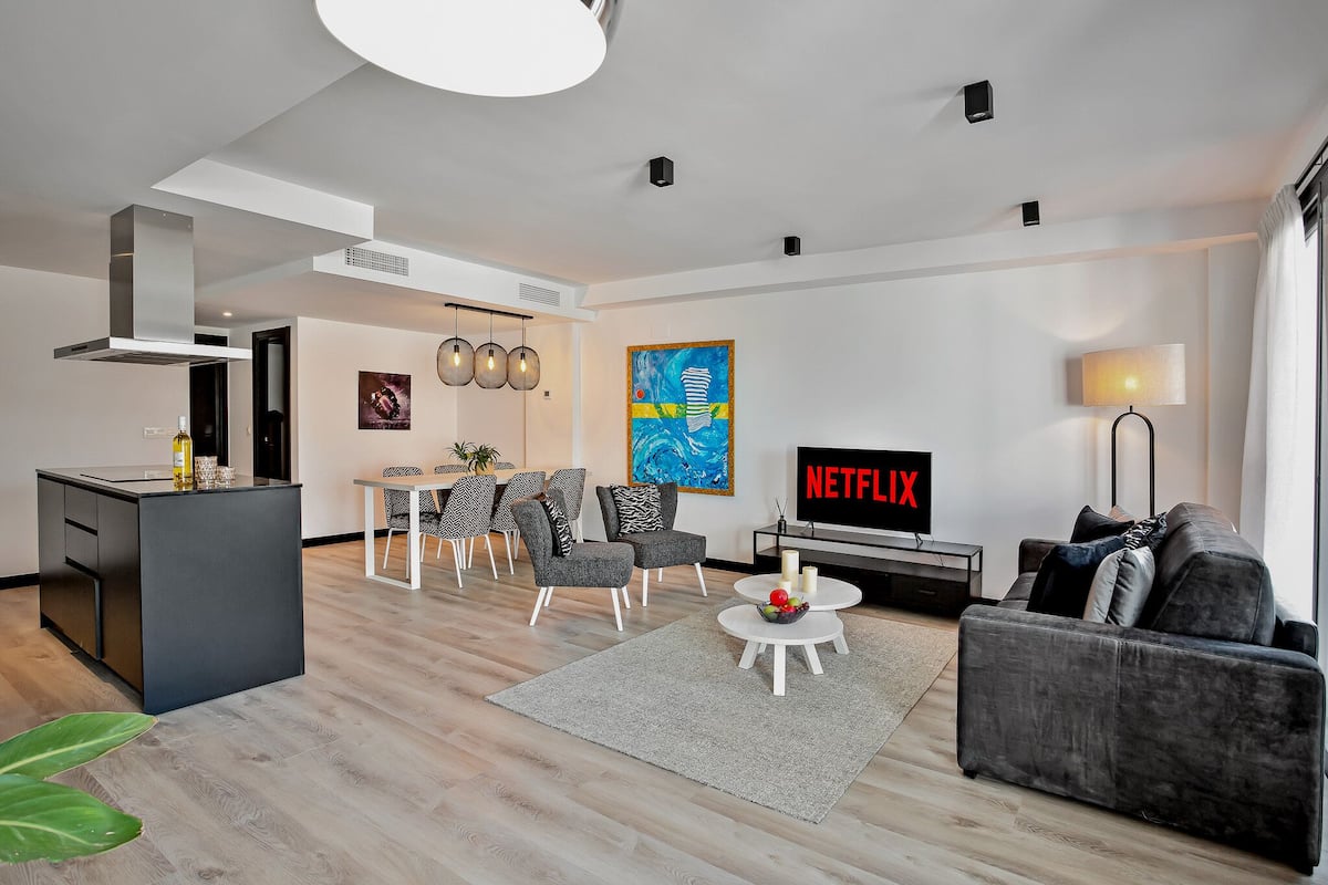 A modern living room with a grey sofa, a dining area with chairs, a kitchen island, and a TV displaying 'NETFLIX'.