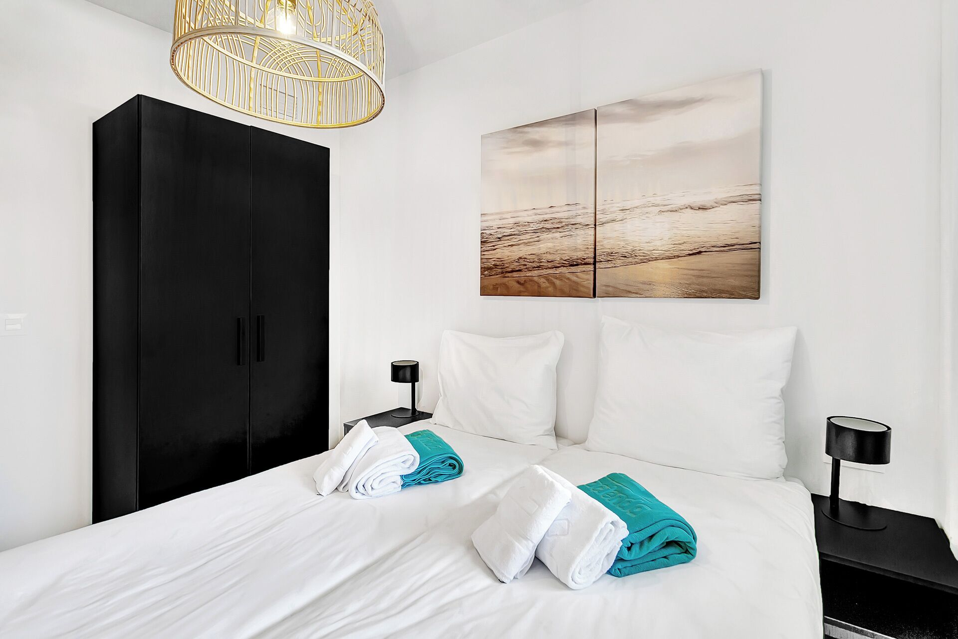 Une chambre d’hôtel avec un grand lit, du linge de lit blanc, une armoire noire, une œuvre d’art accrochée au mur et deux tables de chevet avec des lampes.