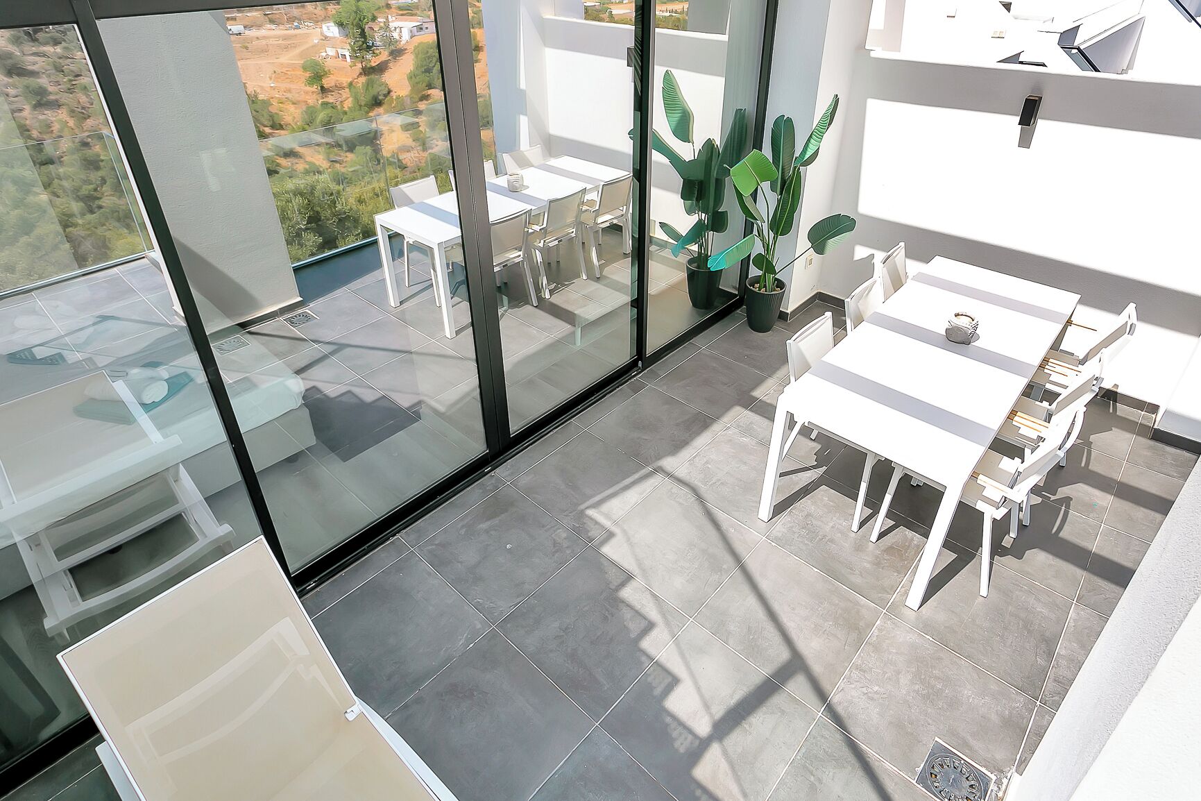 Een balkon met een glazen wand, wit meubilair en plantenpotten.
