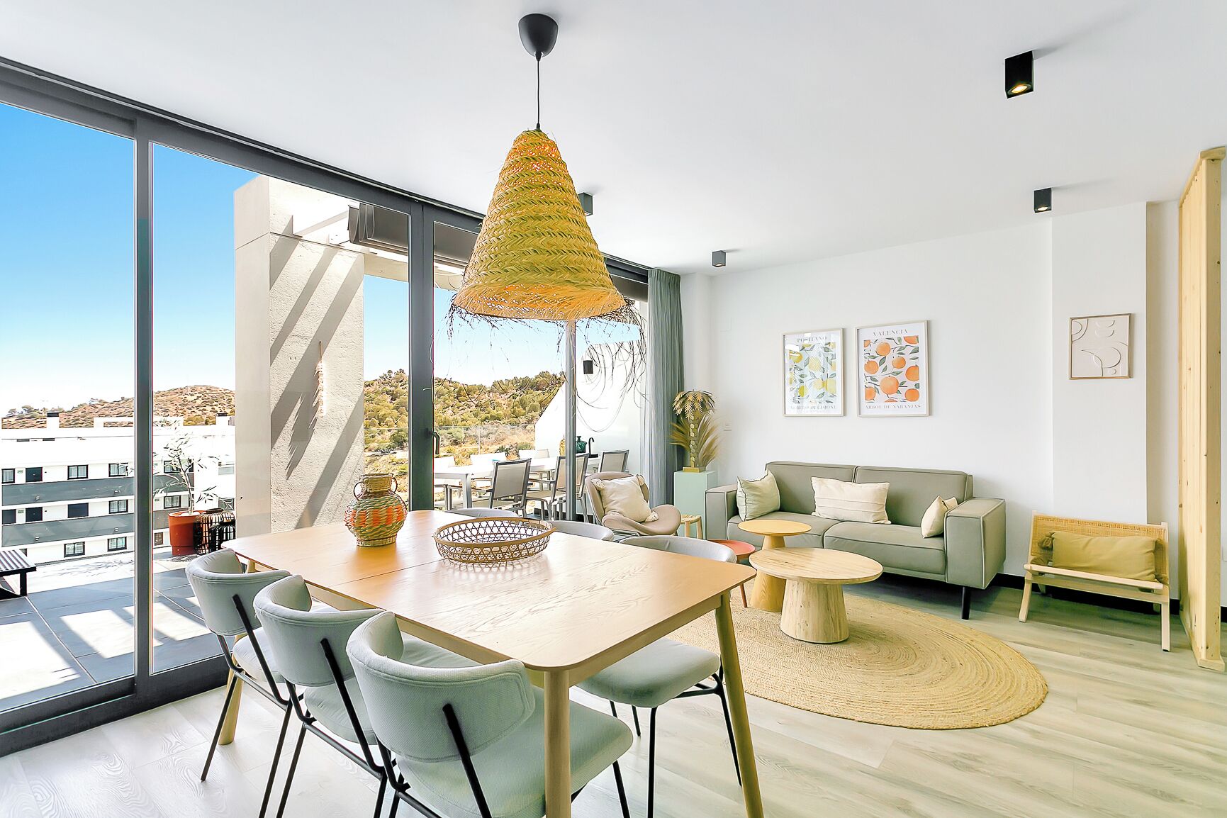 A modern living room with a large window, a dining area with a wooden table and chairs, a sofa, and a hanging light fixture.