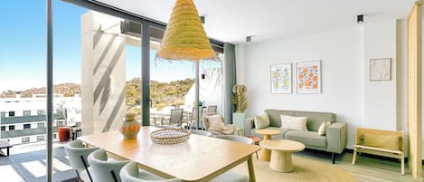 A modern living room with a large window, a dining area with a wooden table and chairs, a sofa, and a hanging light fixture.