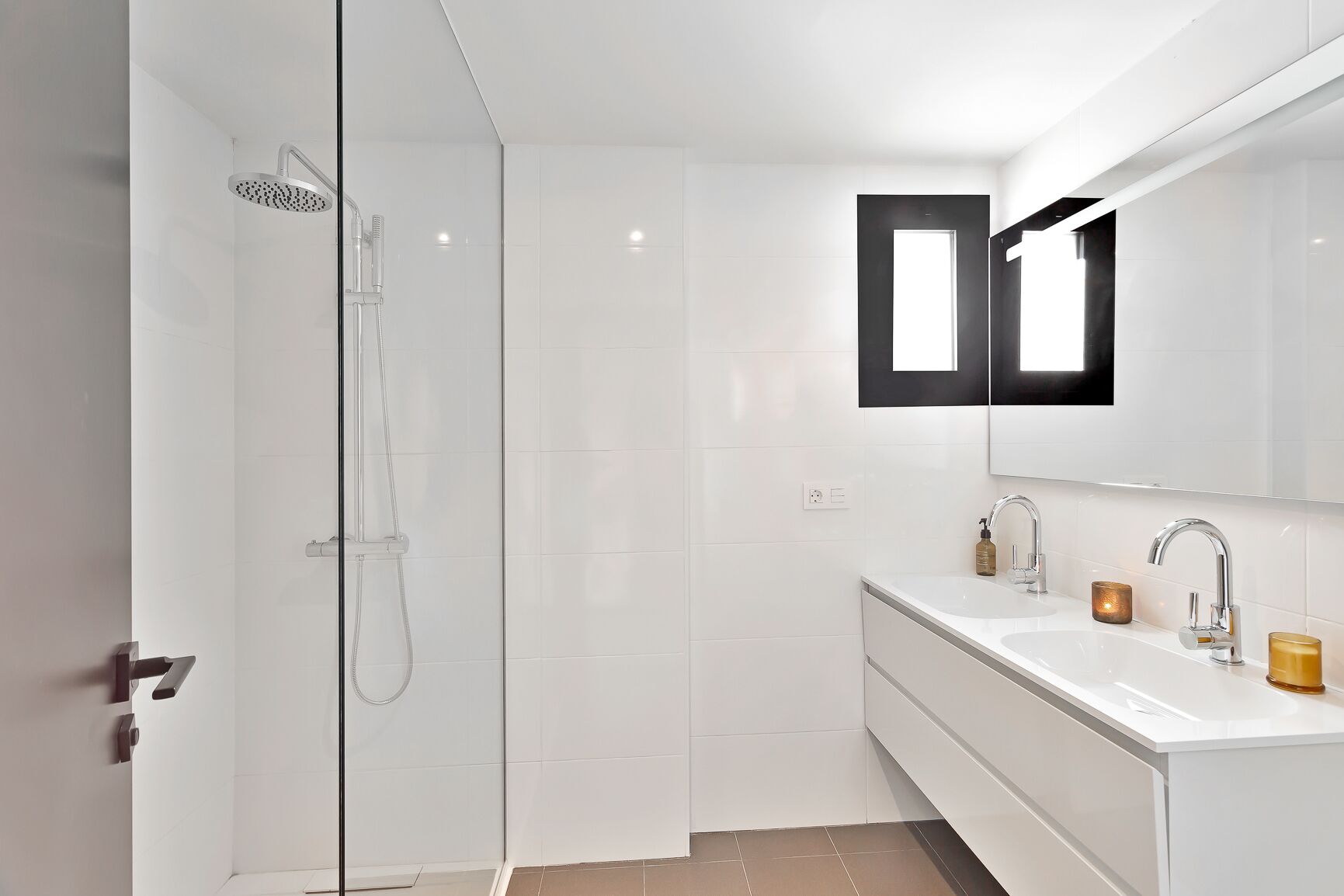 Une salle de bain moderne dotée d’une douche à parois de verre, d’un grand miroir et d’un meuble-lavabo blanc avec deux vasques.