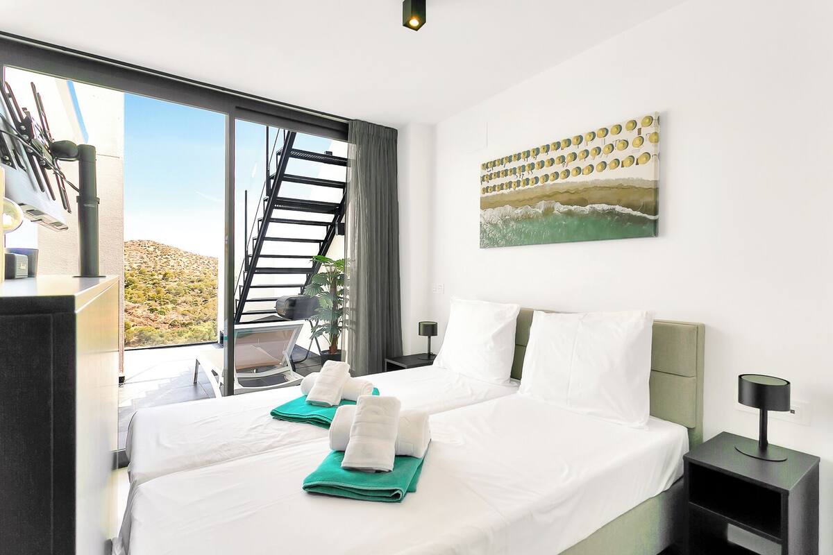 A hotel room with a large bed, a view of the desert through a sliding door, a painting on the wall, and a small table with a lamp.