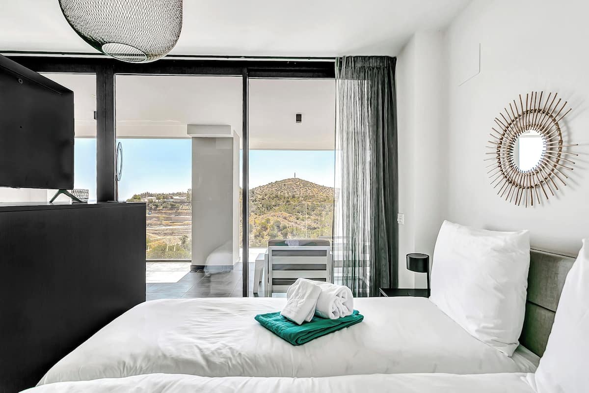 A hotel room with a large bed, a view of a hill, and a balcony with a chair.