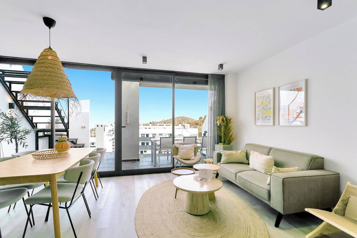 A modern living room with a sofa, coffee table, and dining area. There is a large window with a view of buildings and a staircase.