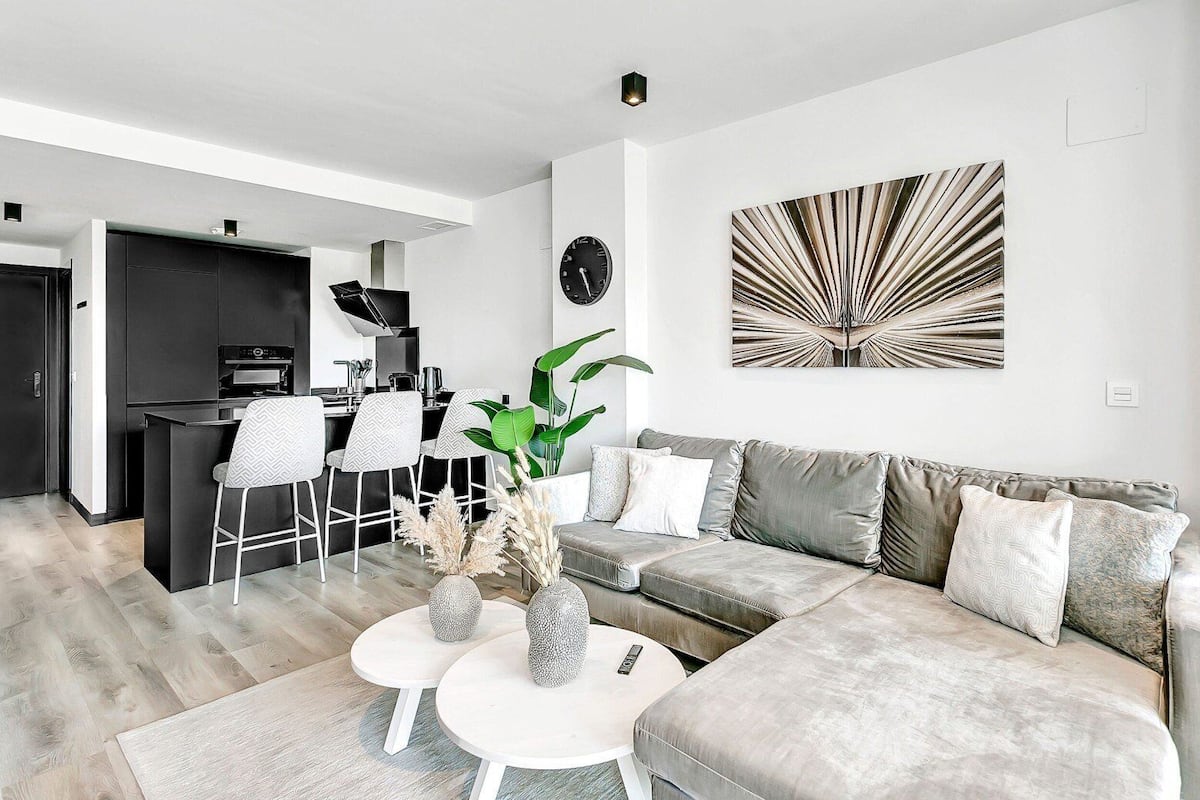 A modern living room with a sofa, two small tables, a coffee table, a dining area with chairs, a kitchen, and a large abstract painting on the wall.