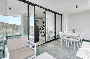 Un salón moderno con un ventanal corredera de vidrio que da a un balcón, una zona de comedor con mobiliario blanco y vistas a un paisaje montañoso.