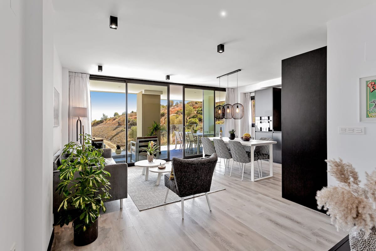 A modern living room with a large sliding glass door leading to a balcony, a dining area with a white table and chairs, and a kitchen area with a black cabinet and pendant lights.