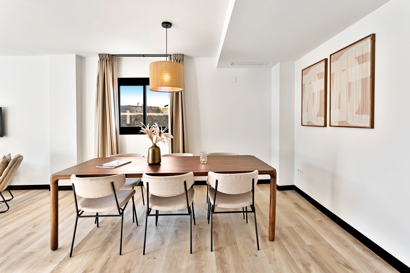 Une salle à manger moderne avec une table en bois, quatre chaises, un vase contenant des fleurs séchées et une grande fenêtre avec des rideaux.