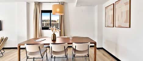 A modern dining room with a wooden table, four chairs, a vase with dried flowers, and a large window with curtains.