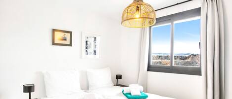 Une chambre d’hôtel avec deux lits, une grande fenêtre donnant sur le paysage, un luminaire suspendu et une petite table avec une lampe.