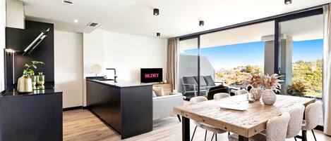 A modern hotel room with a kitchen area, a dining table with chairs, a TV, and a large window offering a view of the outdoors.