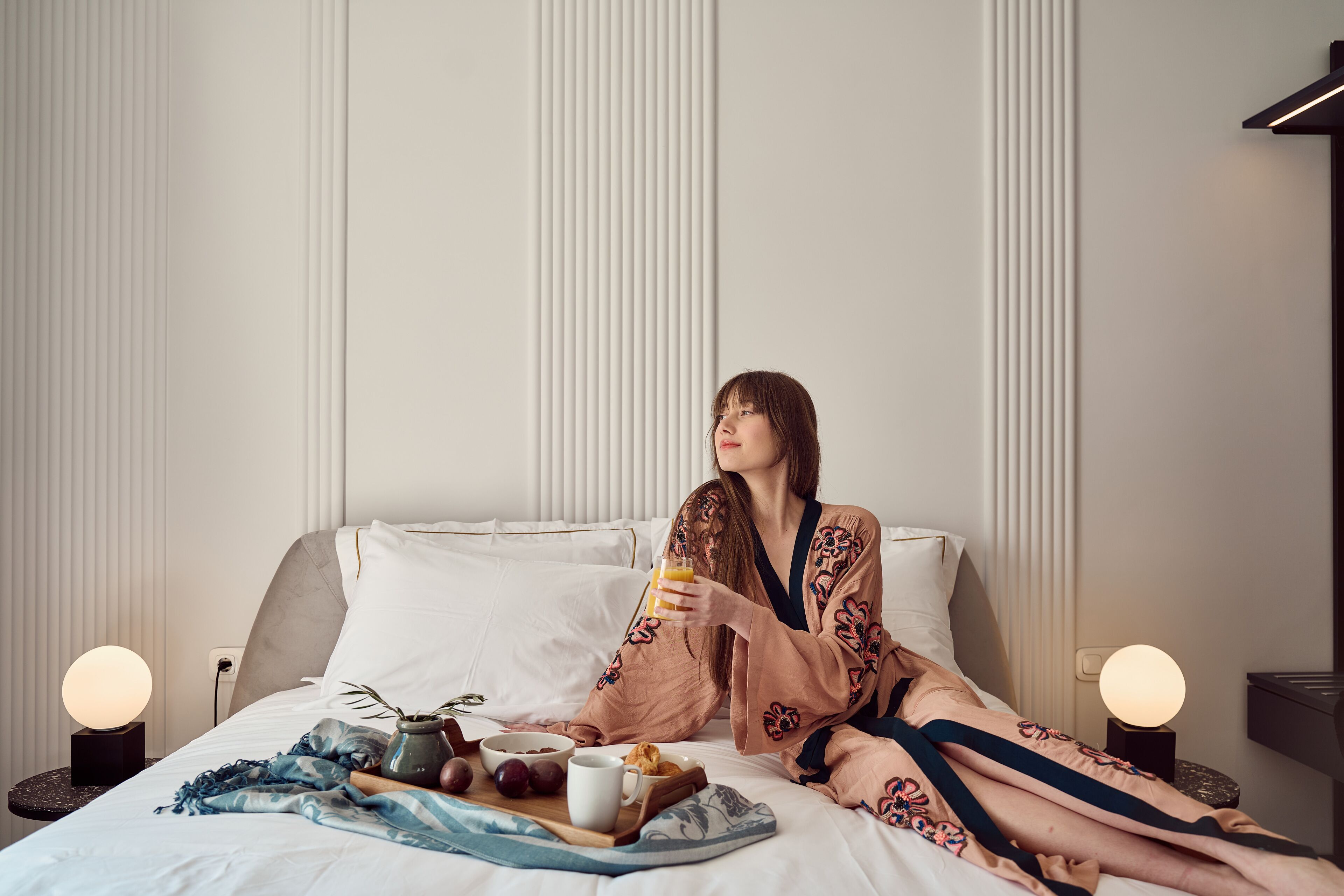 Une personne est assise sur un lit, avec un plateau contenant divers éléments pour le petit-déjeuner, notamment un bol, une tasse et une assiette avec des fruits et des viennoiseries.