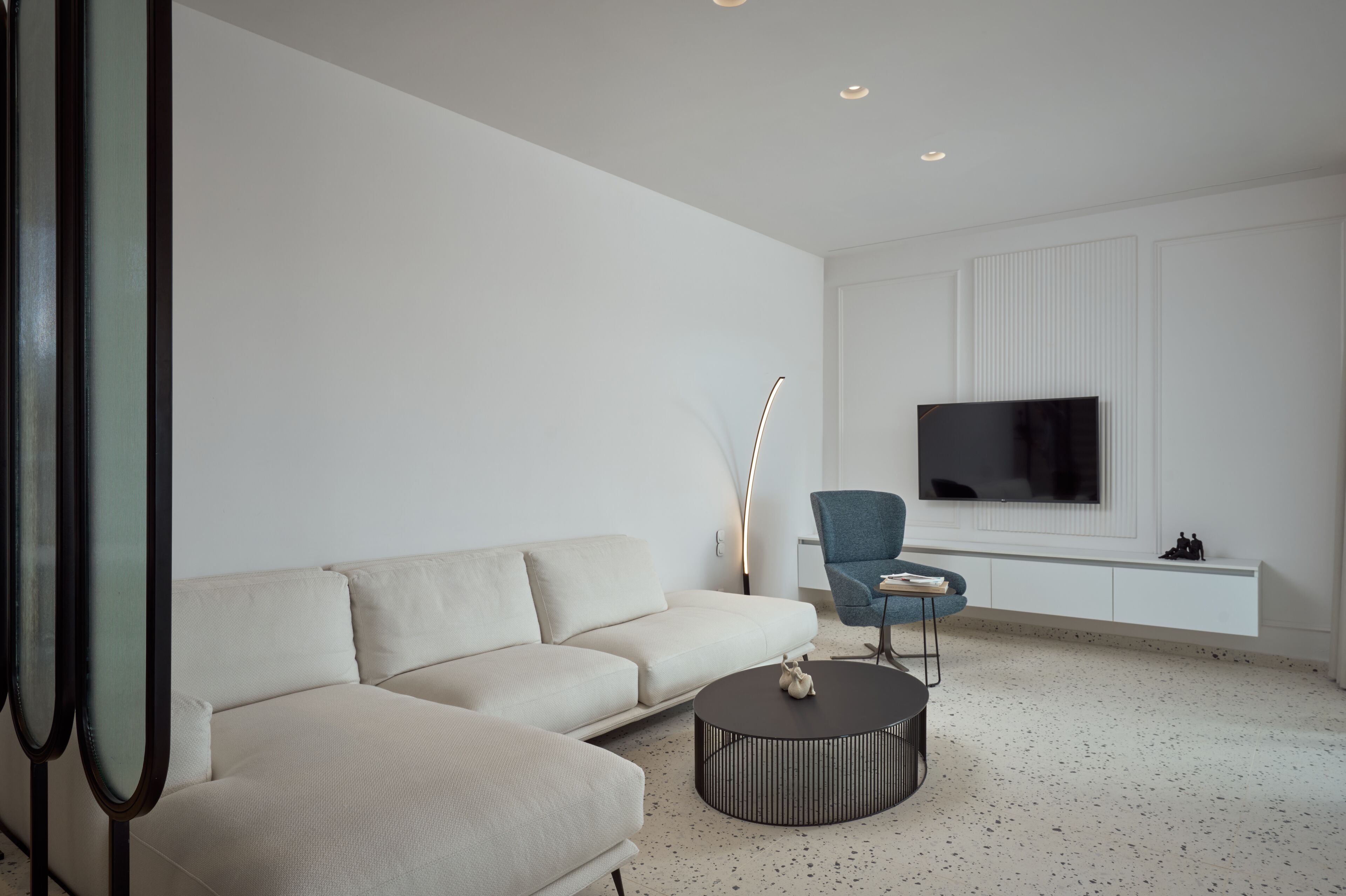 A modern living room with a beige sofa, a round coffee table, a blue armchair, and a flat-screen TV mounted on the wall.