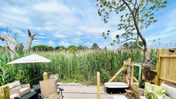An outdoor deck with a wooden bench, a table, and a sofa. There is a large umbrella and a wooden fence with a gate. - 🌾Welcoming Tiny Home! Downtown & Beachy☀️ (Guilford)