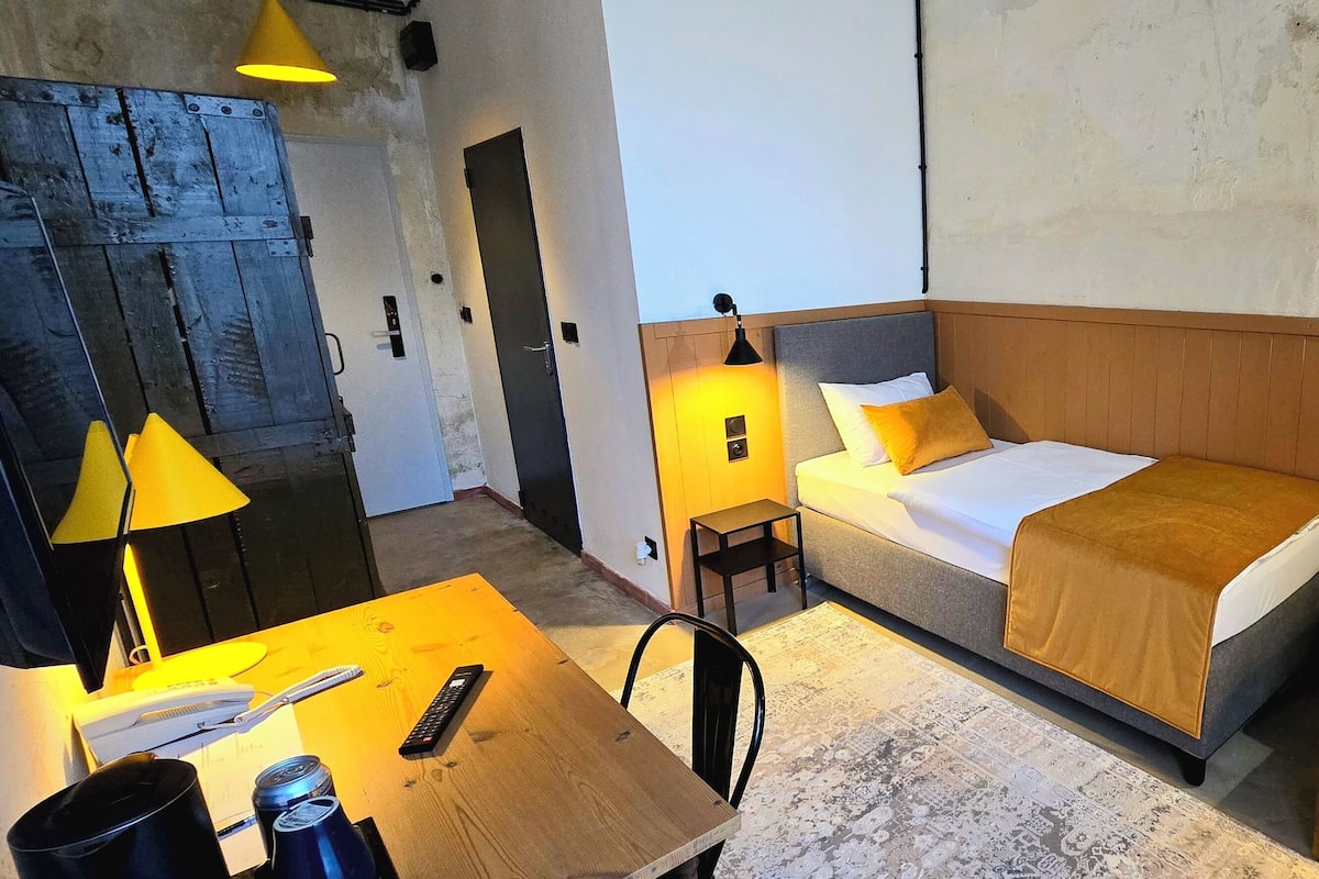 Ein kleines Hotelzimmer mit einem Einzelbett, einem kleinen Tisch mit Lampe, einem Sessel und einer Couch.