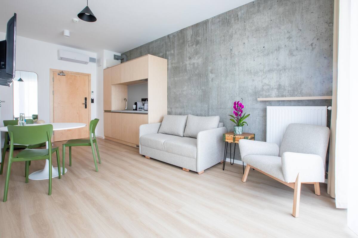 Ein modernes Wohnzimmer mit einer Couch, zwei Sesseln, einem Esstisch mit Stühlen und einer Küchenzeile.