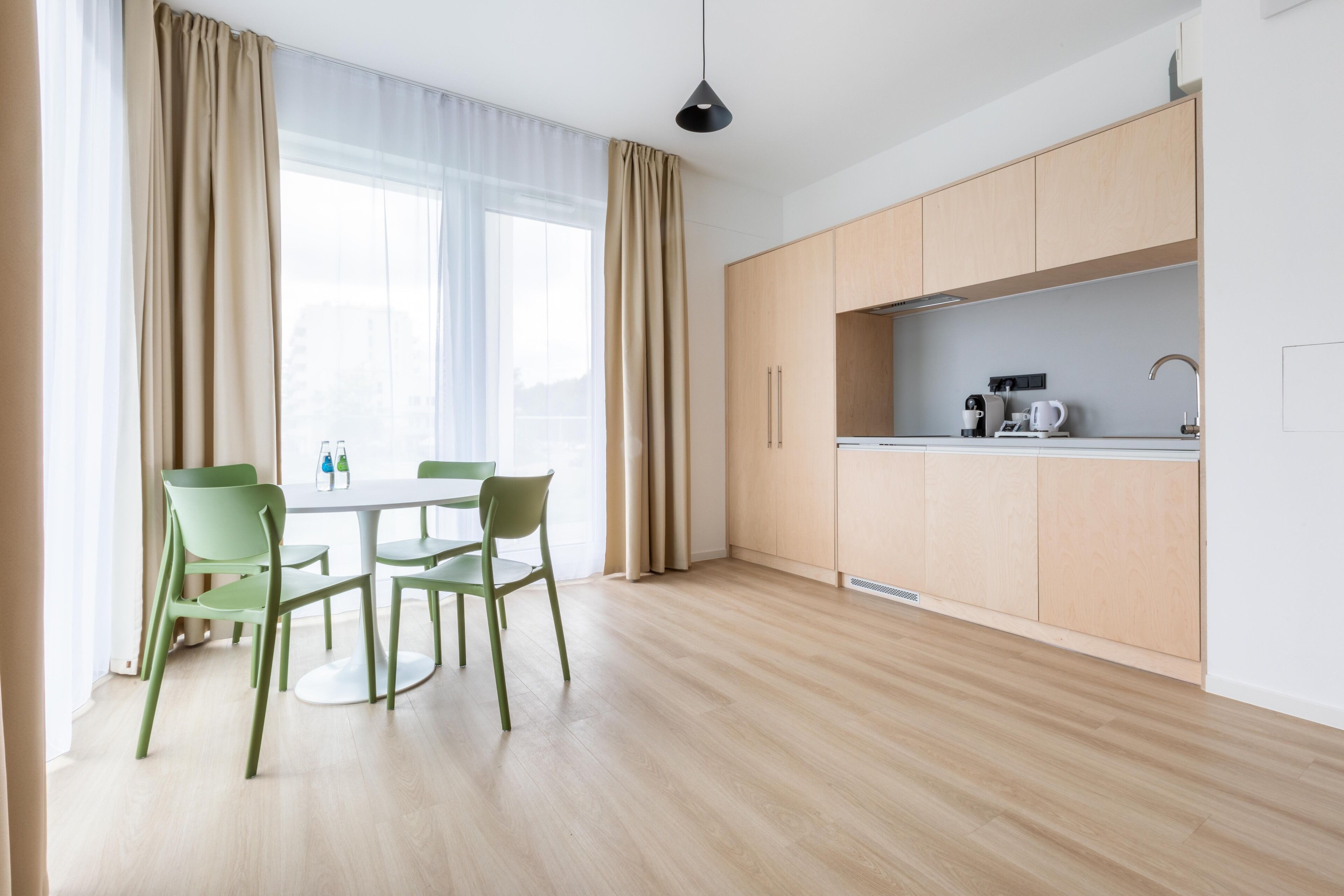 A modern kitchen with light wooden cabinets, a white countertop, and a sink. There is a dining area with a round table and green chairs.
