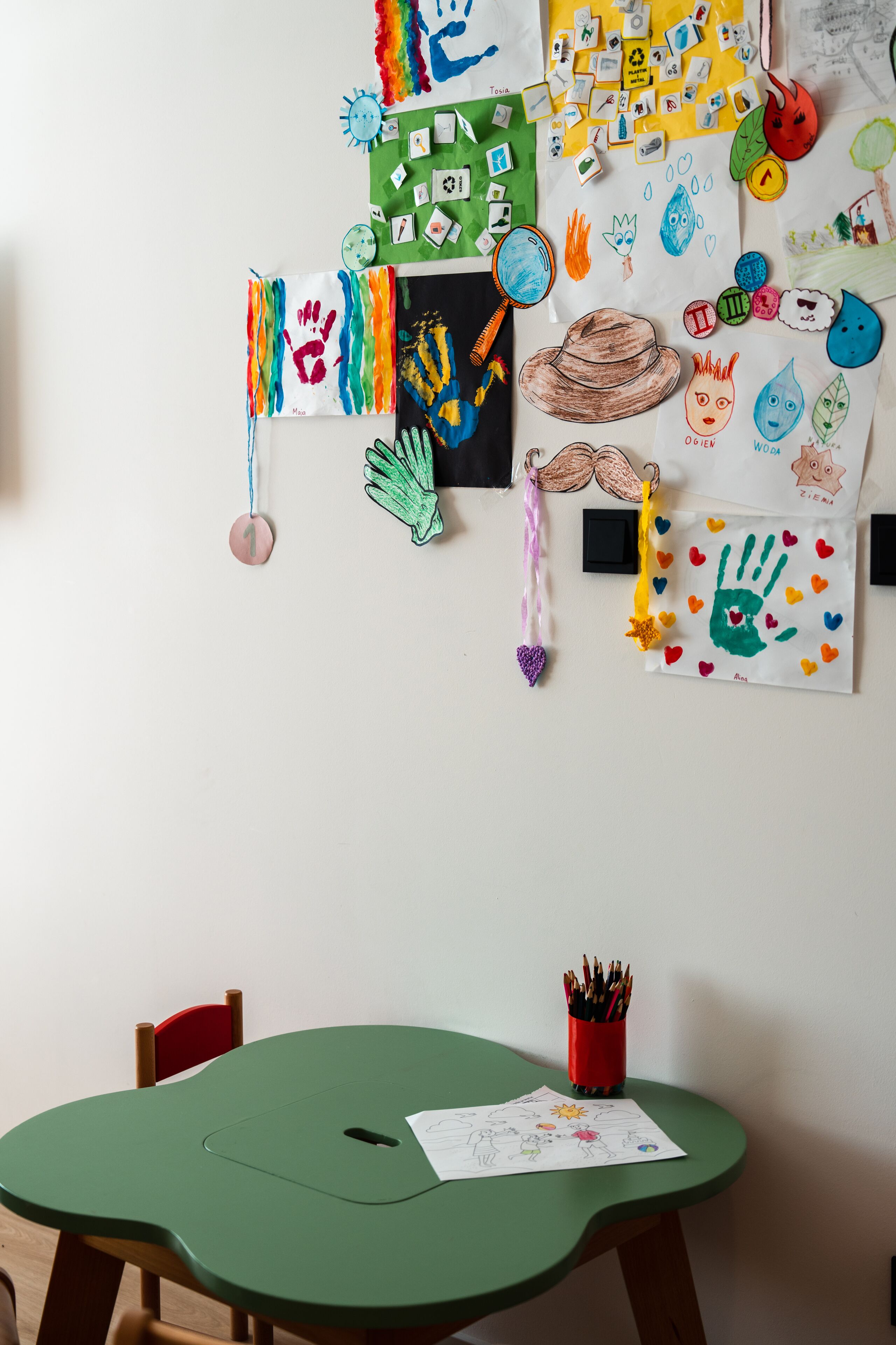 A child's art project on a wall, a green table with a chair, and a red pencil holder with pencils.