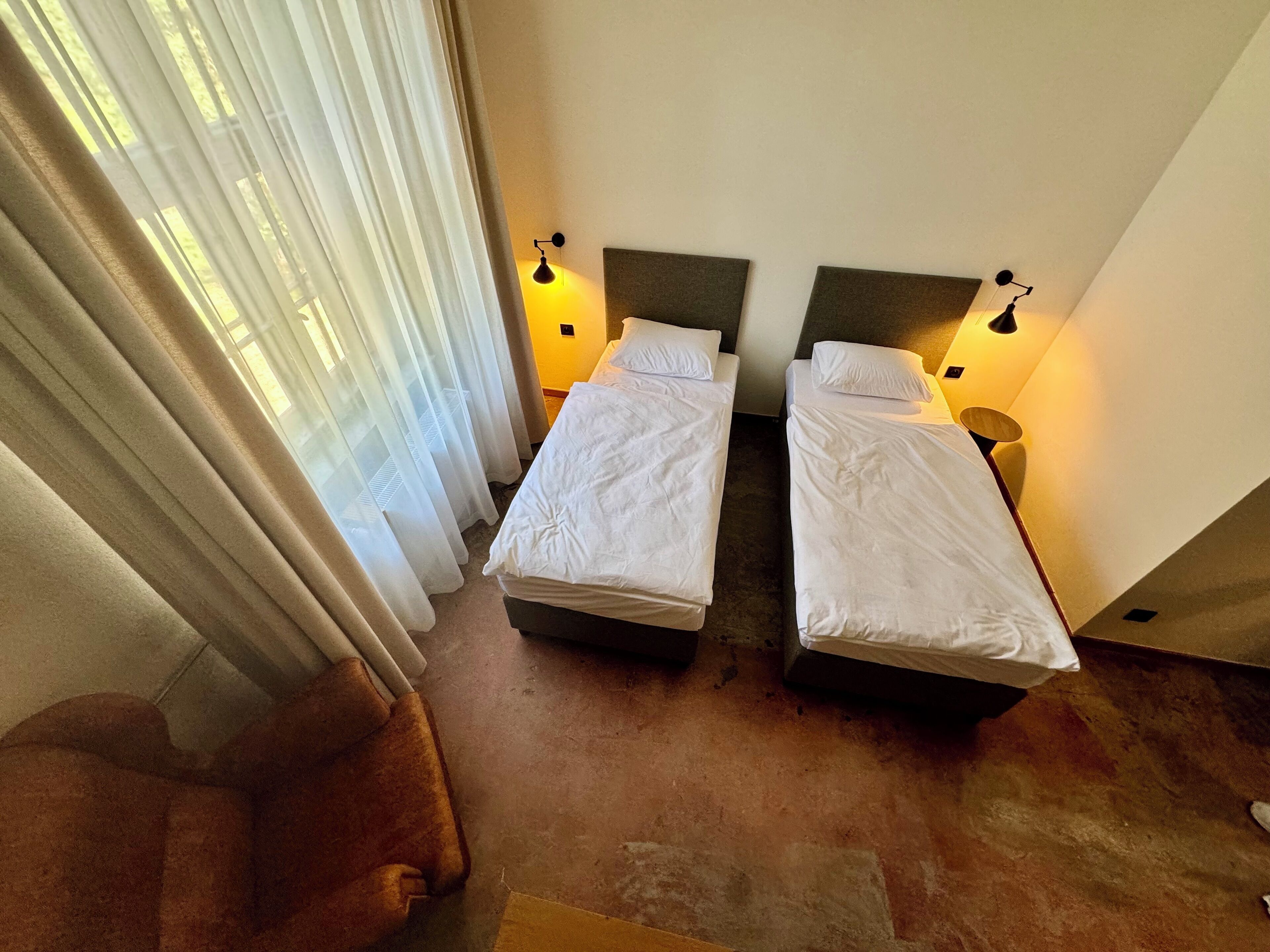 A room with two beds, each with a pillow and a nightstand. There is a chair and a window with curtains.