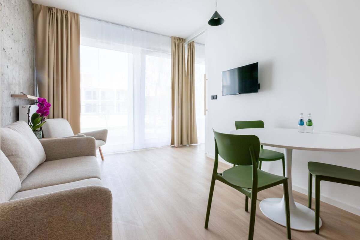 Ein modernes Wohnzimmer mit einer Couch, einem Esstisch mit Stühlen, einem an der Wand montierten Flachbildfernseher und einem großen Fenster mit Vorhängen.