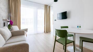 Una sala de estar moderna con un sofá, una mesa de comedor con sillas, un televisor de pantalla plana montado en la pared y un amplio ventanal con cortinas.