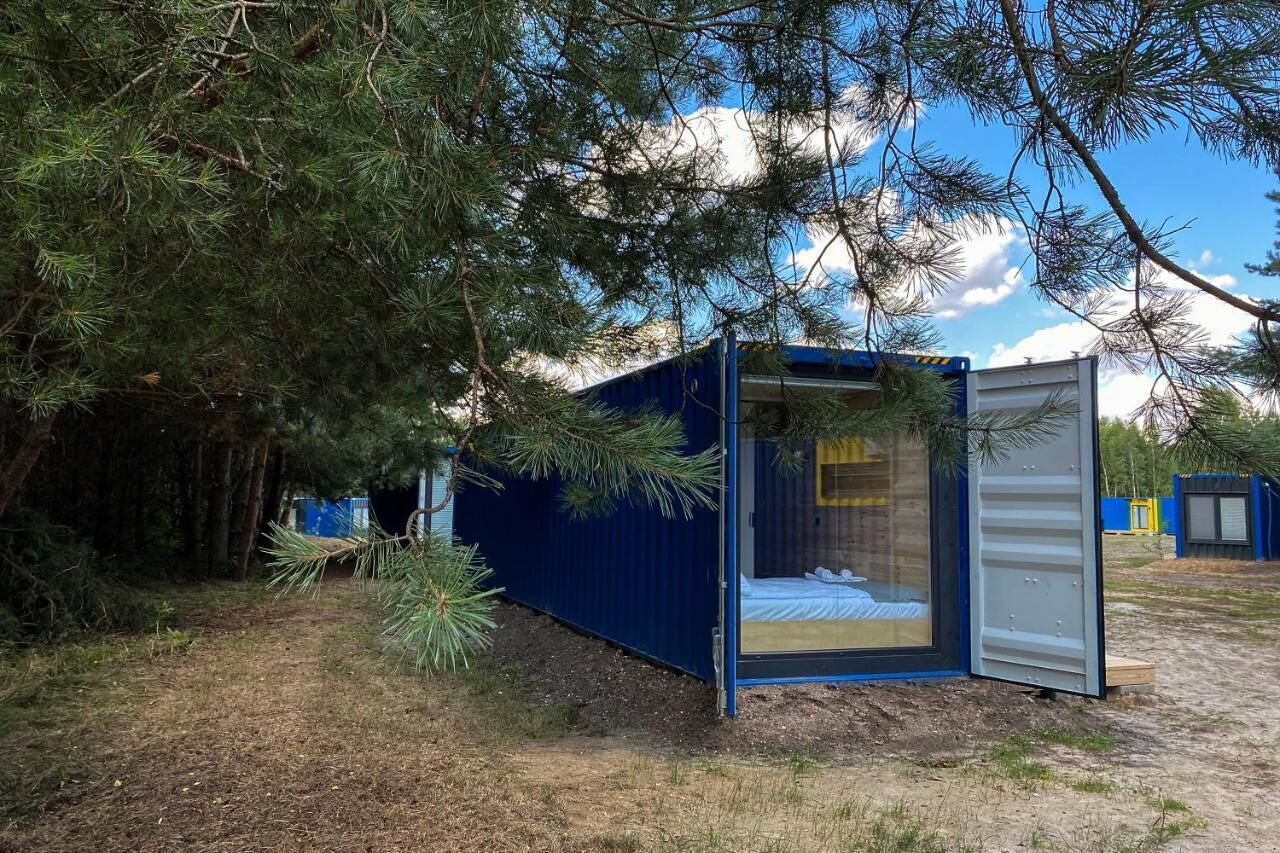 A small, blue, container-style cabin with a bed and a window, surrounded by trees.