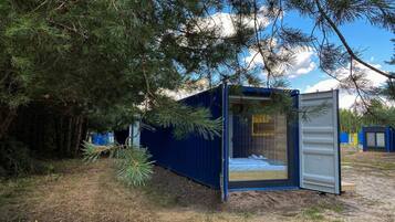 A small, blue, container-style cabin with a bed and a window, surrounded by trees.
