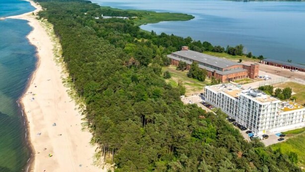 Vue aérienne d’une propriété située sur la côte, avec une plage, une forêt dense et un grand bâtiment à étages.