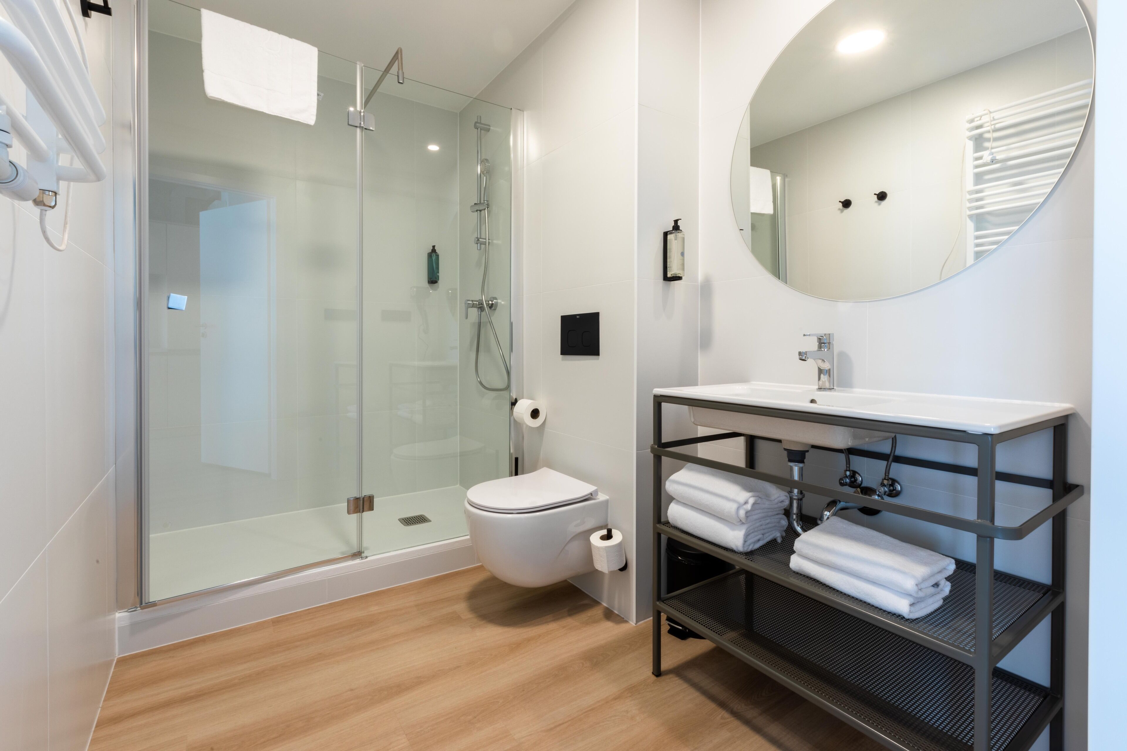 A modern bathroom with a glass-enclosed shower, a white toilet, a sink with a mirror, and a metal shelf with towels.