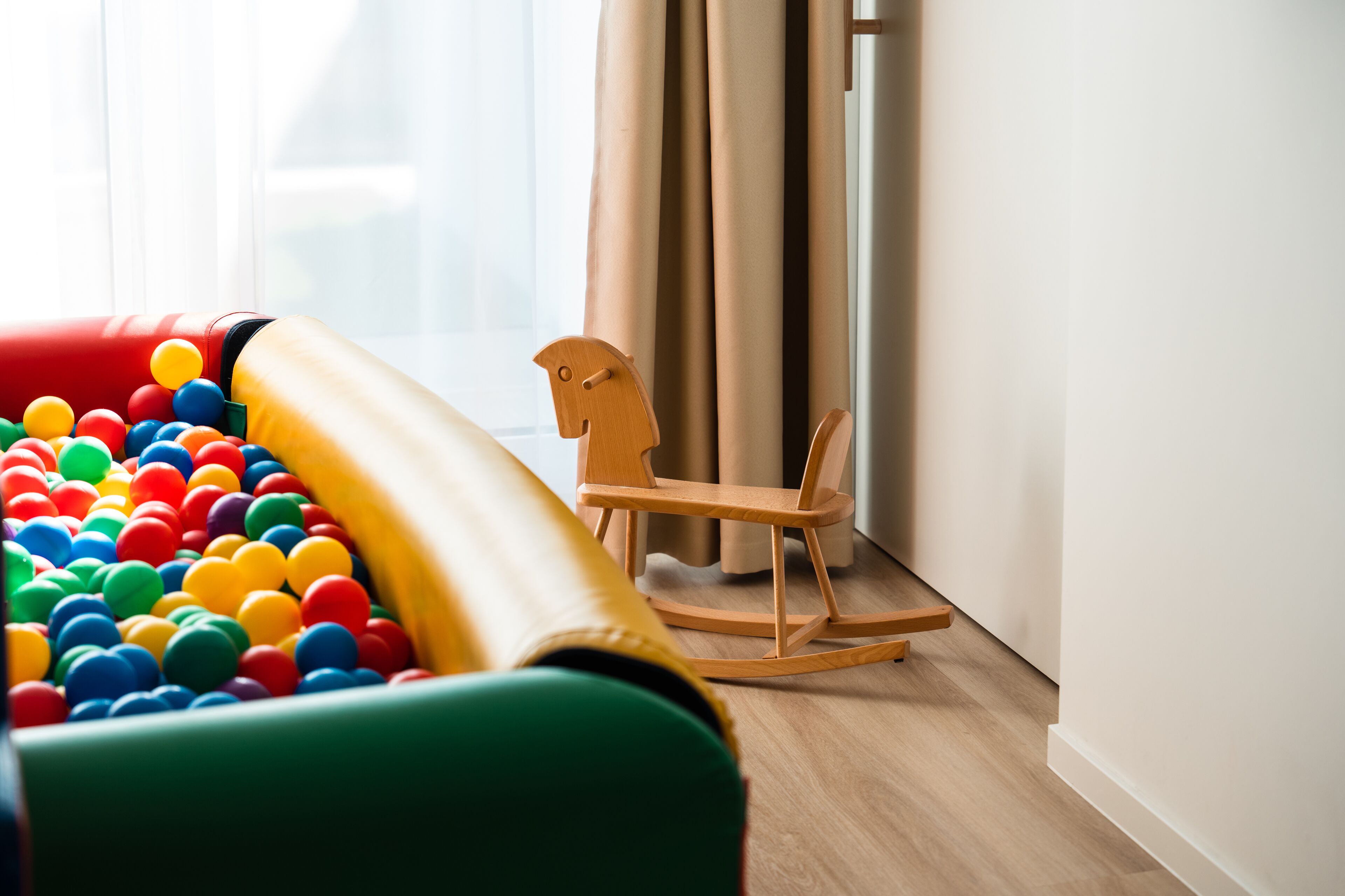 Een kamer met een kleurrijk knikkerbad, een houten schommelstoel en een raam met gordijnen.