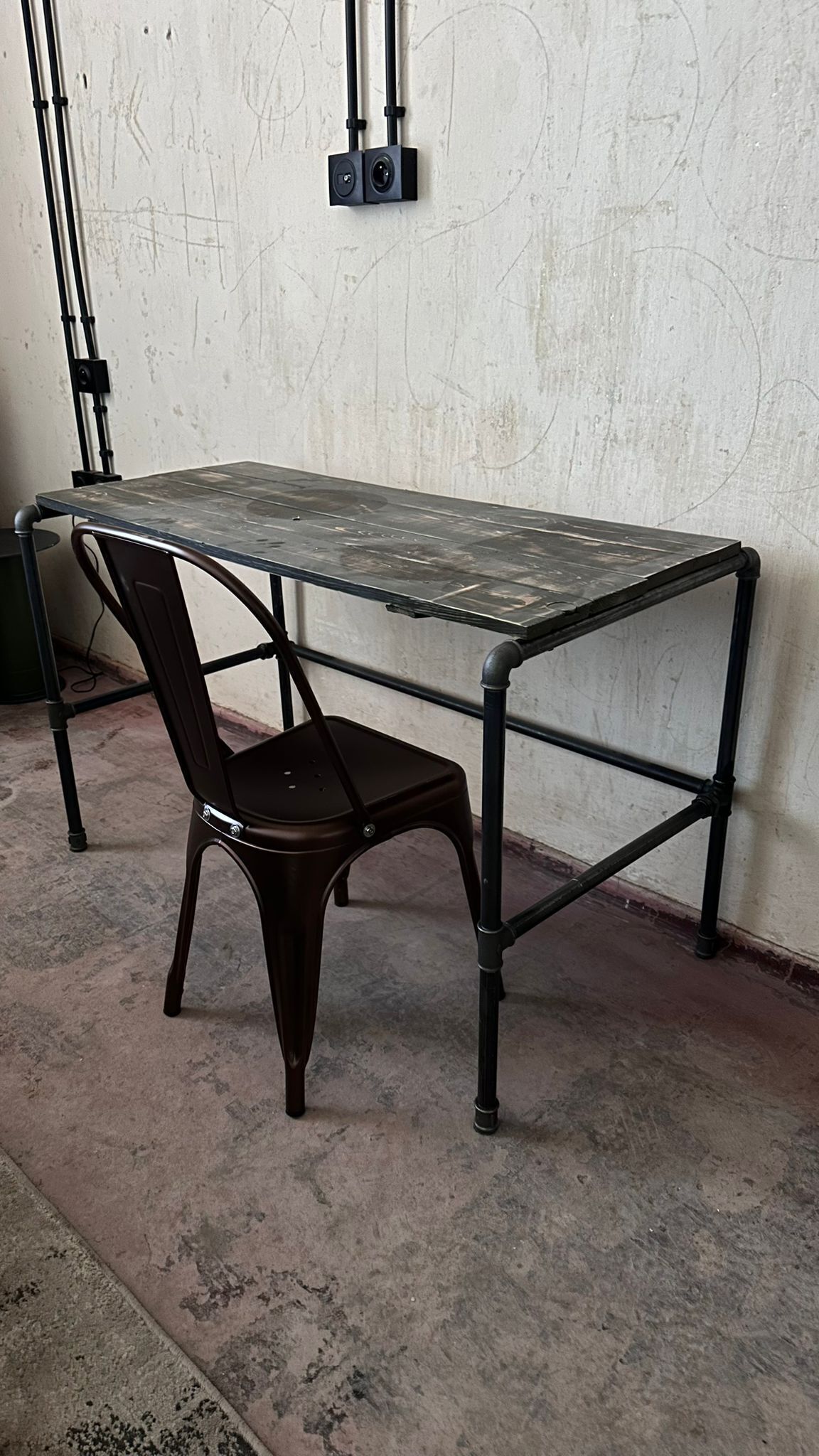 Une table à structure métallique, accompagnée d’une chaise, se trouve dans une pièce dont le sol est en béton et dont le mur est uni.