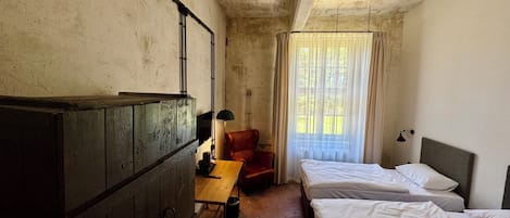 A room with two beds, a wooden desk, a chair, a lamp, and a window with curtains.