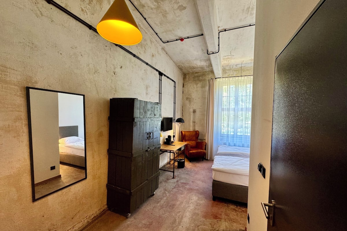 Ein kleines Hotelzimmer mit einem großen Spiegel, einem Kleiderschrank, einem Schreibtisch mit Stuhl, einem Bett und einer Hängeleuchte.