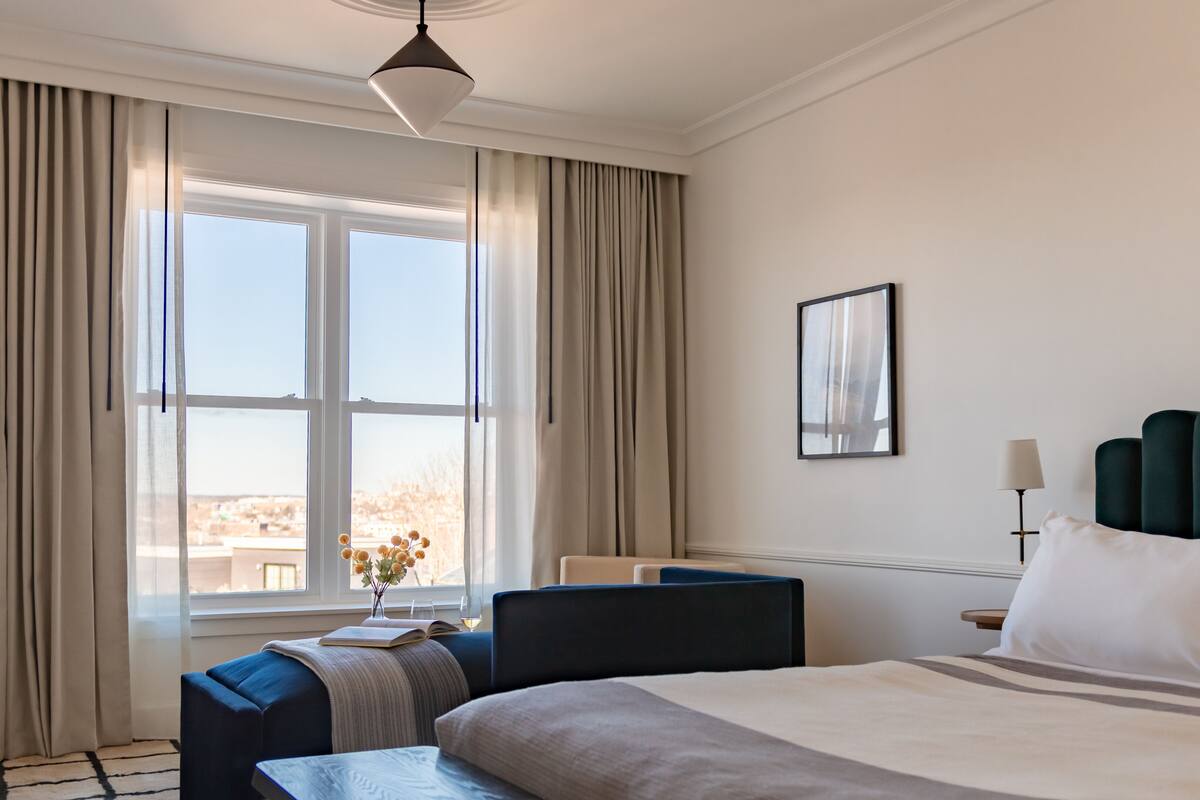 Ein Hotelzimmer mit einem großen Bett, einem Nachttisch, einem Sessel, einem Schreibtisch, einer Lampe und einem fenster mit Vorhängen.