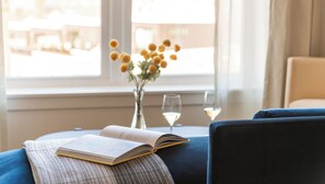 A cozy reading nook with a book, a vase with flowers, and two glasses of white wine on a table by a window.